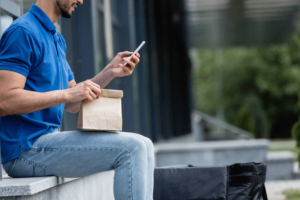 Cropped view of smiling courier using smartphone and holding paper bag outdoors  - 写真・画像