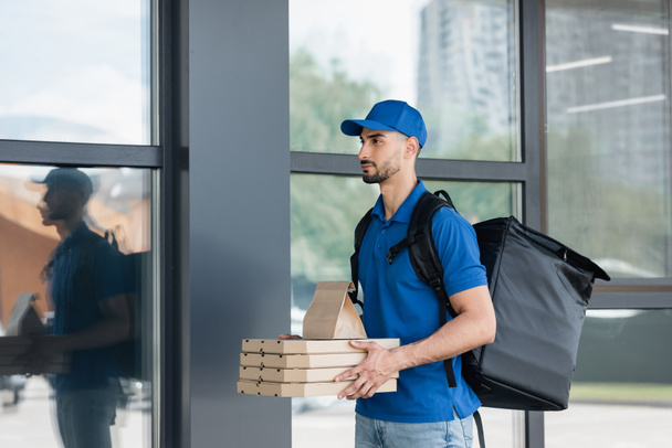Arabian deliveryman holding paper bag and pizza boxes near building  - Foto, imagen