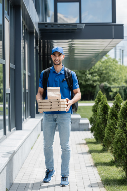 Positive muslim courier holding paper bag and pizza boxes near building  - Fotoğraf, Görsel