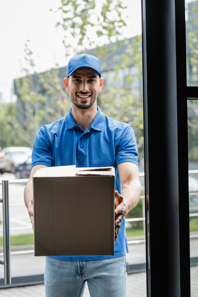Smiling muslim courier holding carton box near door  - Foto, imagen
