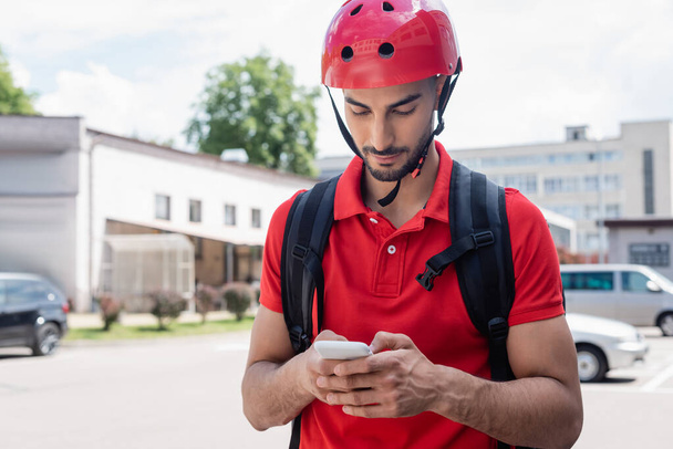 Arabian courier in helmet using smartphone on urban street  - 写真・画像
