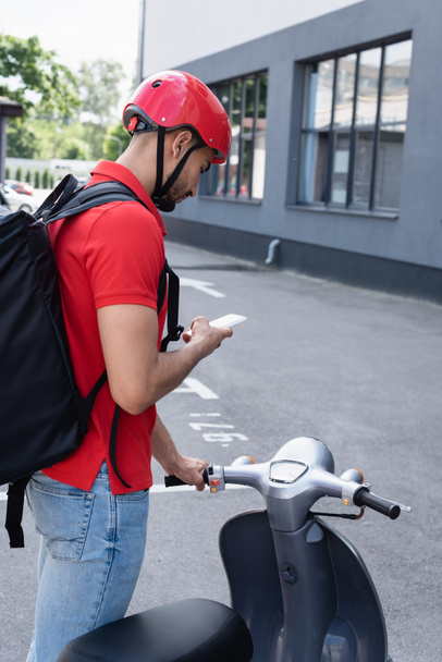 Arabisch deliveryan in helm met behulp van smartphone in de buurt van scooter buiten  - Foto, afbeelding