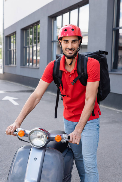Smiling muslim courier with thermo backpack standing near scooter  - Photo, Image