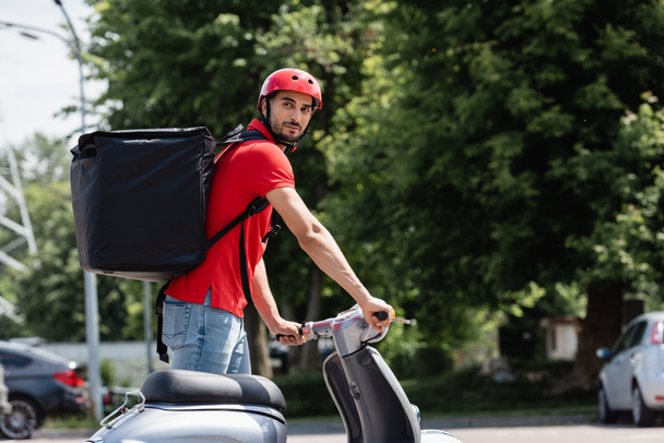 Young arabian deliveryman with thermo backpack standing near scooter on urban street  - 写真・画像