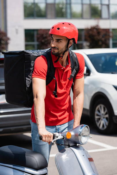 Arabian deliveryman in helmet and thermo backpack standing near scooter outdoors  - Fotó, kép