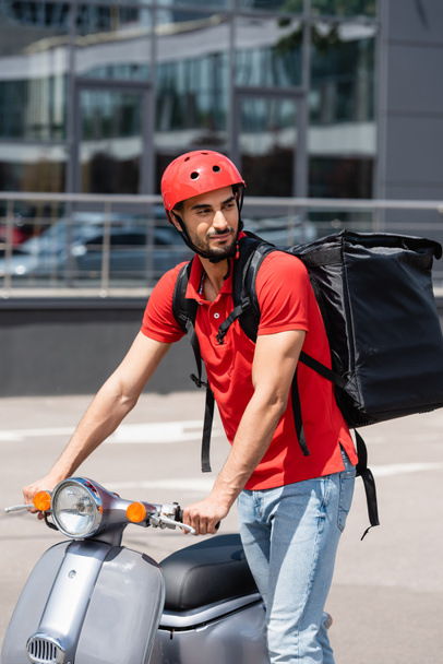 Arabian deliveryman in helmet and thermo backpack standing near scooter outdoors  - 写真・画像