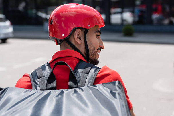 Vista lateral del repartidor musulmán en casco y mochila térmica al aire libre  - Foto, imagen