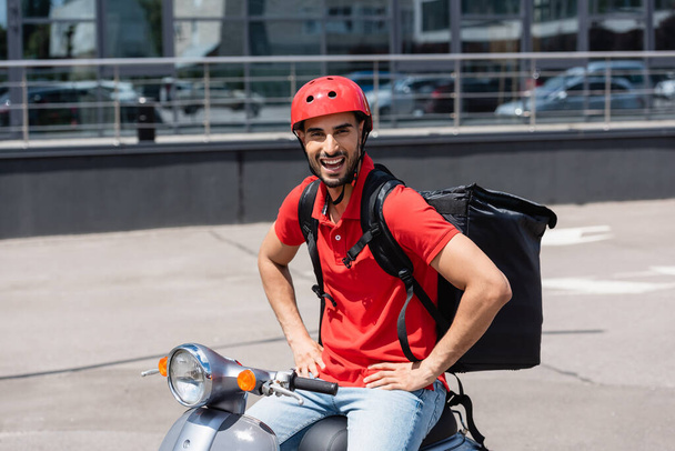 Excited arabian deliveryman with thermo backpack holding hands on hips while sitting on scooter  - Foto, immagini