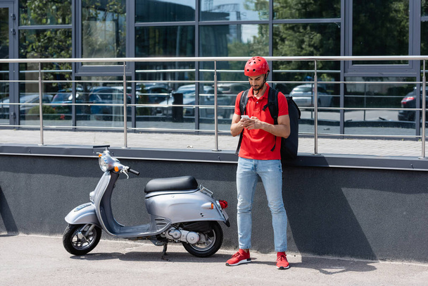 Young arabian deliveryman with thermo backpack using smartphone near scooter and building outdoors  - Фото, изображение