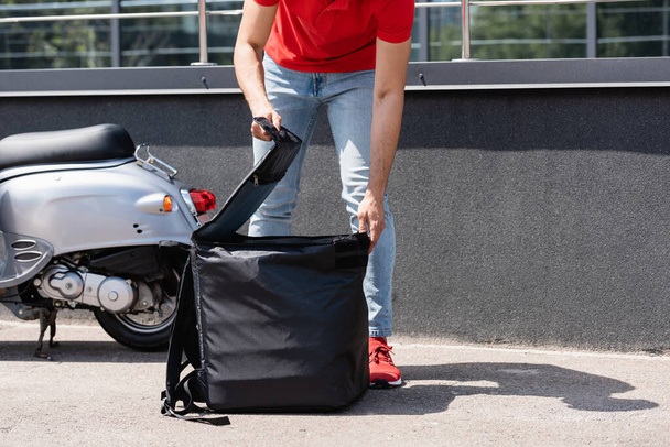Cropped view of deliveryman opening thermo backpack near scooter  - Foto, Imagem