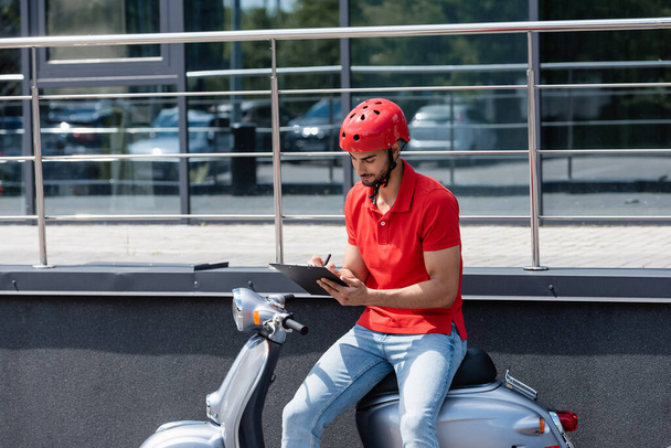 Young muslim courier in helmet writing on clipboard while sitting on scooter outdoors  - Foto, afbeelding