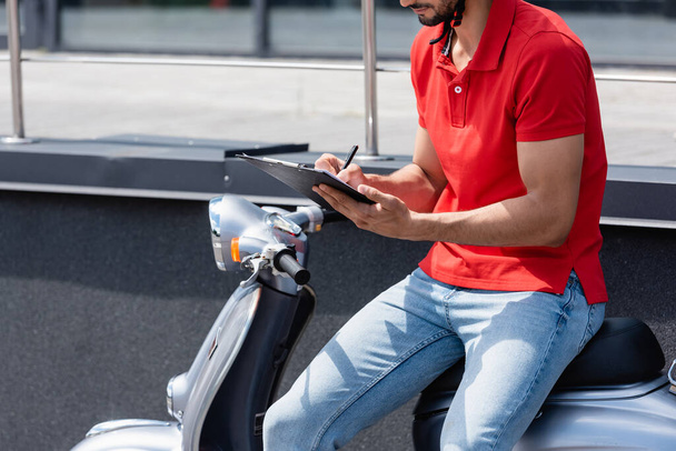 Cropped view of courier writing on clipboard on scooter on urban street  - 写真・画像