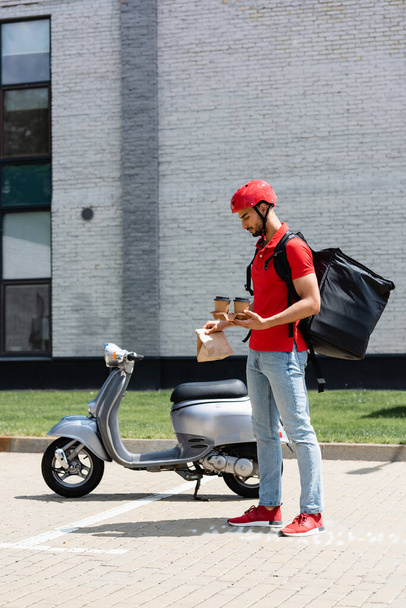 Side view of arabian courier with thermo backpack holding coffee to go and paper bag near scooter  - Photo, image
