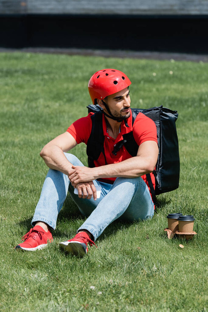 Junger muslimischer Kurier mit Helm und Thermo-Rucksack hält Smartphone in der Nähe von Pappbechern auf Gras  - Foto, Bild