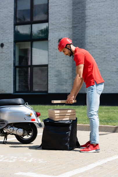 Side view of arabian courier in helmet holding pizza box near thermo backpack and scooter  - Fotografie, Obrázek
