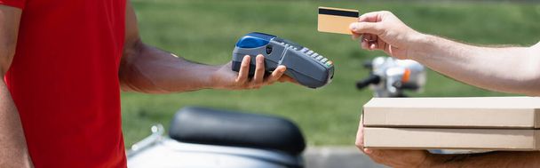 Vista recortada del hombre con cajas de pizza con tarjeta de crédito cerca de mensajero con terminal de pago al aire libre, pancarta  - Foto, Imagen