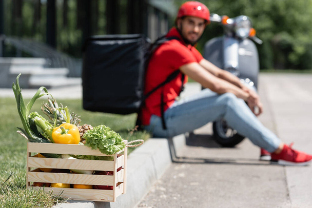 Box with fresh vegetables near blurred deliveryman and scooter outdoors  - 写真・画像
