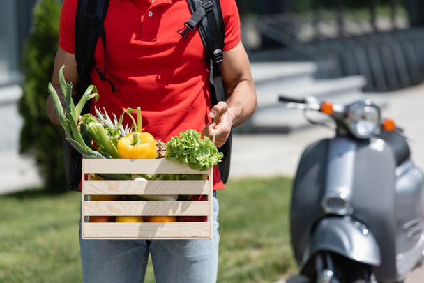 Cropped view of deliveryman in red t-shirt holding box with organic vegetables near blurred scooter  - Fotoğraf, Görsel