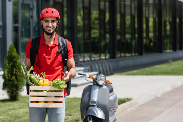 Positive arabian courier holding wooden box with fresh vegetables near blurred scooter  - Foto, Imagen