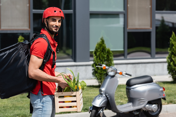 Arabian courier with vegetables and thermo backpack smiling at camera near blurred scooter  - Photo, Image