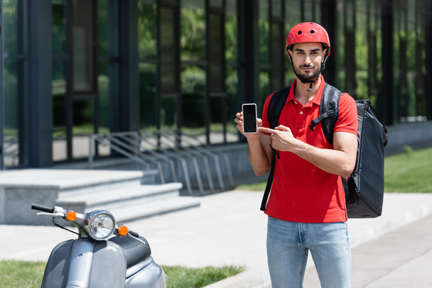 Arabian courier en casco y mochila termo apuntando al teléfono inteligente con pantalla en blanco cerca de scooter al aire libre  - Foto, imagen
