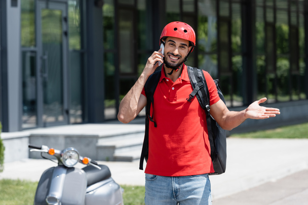Alegre mensajero musulmán con mochila térmica hablando en un teléfono inteligente cerca de scooter borroso al aire libre  - Foto, imagen
