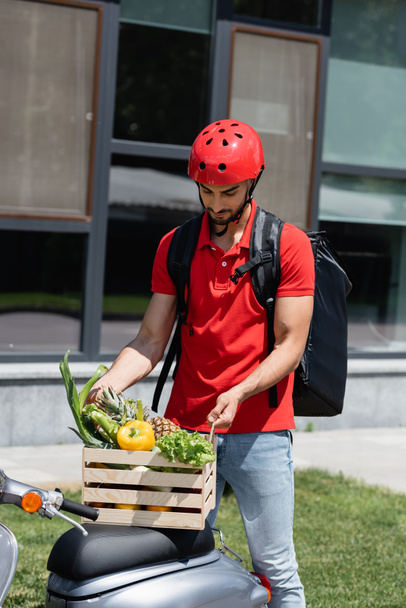 Young arabian courier in safety helmet holding wooden box with vegetables on scooter  - Fotografie, Obrázek