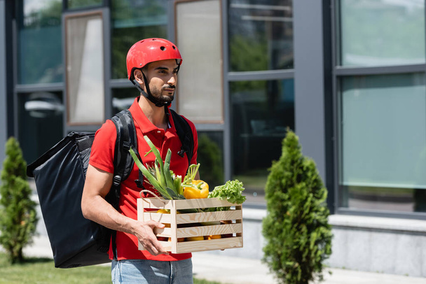 Muslim deliveryman in protective helmet carrying fresh vegetables outdoors  - Фото, изображение