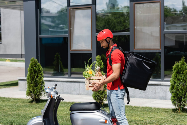 Side view of arabian courier holding box with vegetables near scooter outdoors  - 写真・画像