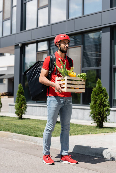 Arabian courier in protective helmet holding box with vegetables outdoors  - Foto, imagen