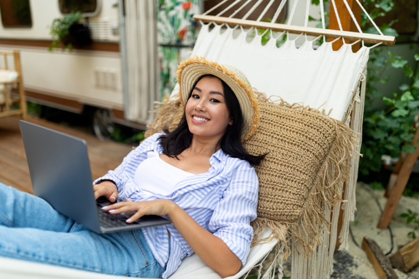 Alegre señora asiática usando el ordenador portátil mientras que miente en la hamaca cerca de RV en viaje que acampa del otoño - Foto, imagen