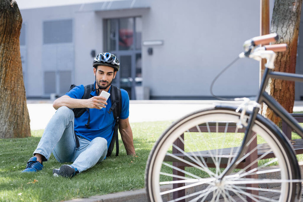 Арабский курьер в шлеме с помощью смартфона на газоне возле размытого велосипеда на городской улице  - Фото, изображение