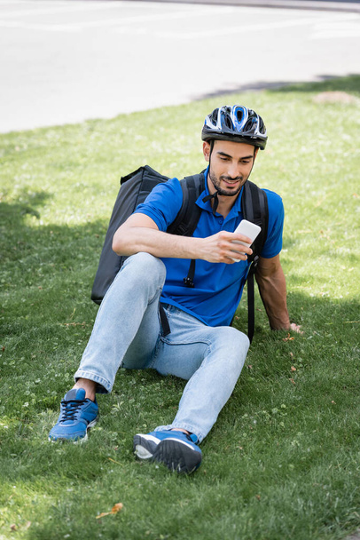 Smiling muslim courier in helmet and thermo backpack using smartphone on lawn  - Fotó, kép