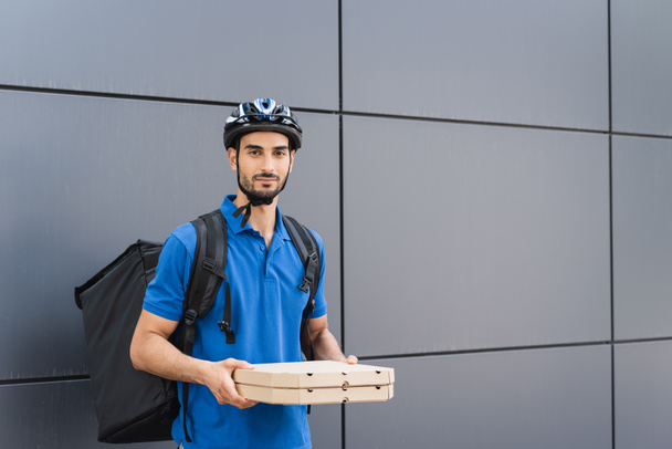 Arabischer Kurier im Helm hält Pizzakartons in der Nähe von Haus im Freien  - Foto, Bild