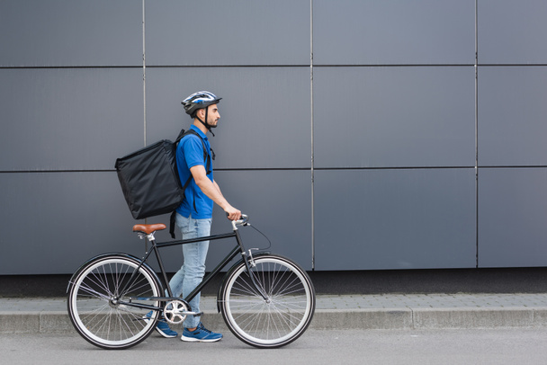 Side view of arabian courier with thermo backpack walking near bike and building outdoors  - Foto, afbeelding