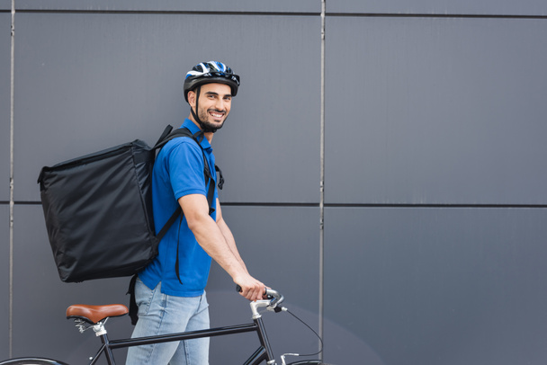 Cheerful muslim deliveryman with backpack and bike walking near building outdoors  - Fotó, kép