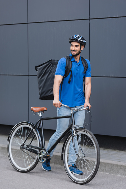 Smiling muslim deliveryman with backpack walking near building on urban street  - Zdjęcie, obraz