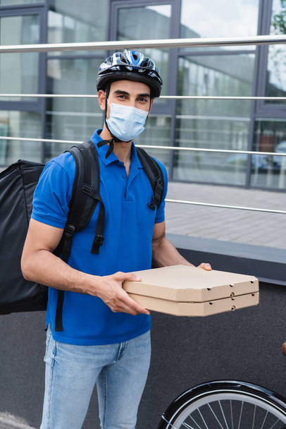 Muslim courier in medical mask holding pizza boxes near bike outdoors  - Φωτογραφία, εικόνα