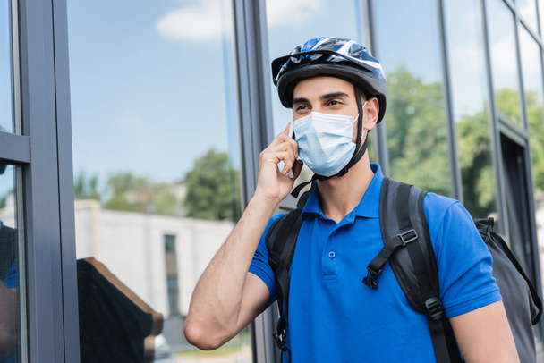 Arabische bezorger met fietshelm en medisch masker in gesprek op smartphone in stedelijke straat  - Foto, afbeelding