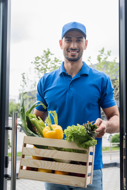 Smiling arabian deliveryman holding box with vegetables near door  - Фото, зображення