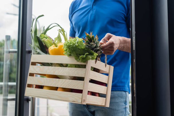 Cropped view of deliveryman holding box with fresh vegetables near door - Photo, Image