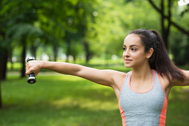 young sportswoman smiling while exercising with dumbbell in park - Foto, Bild