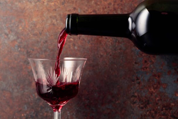 Il vino rosso viene versato da una bottiglia in un bicchiere di vino su sfondo marrone arrugginito.  - Foto, immagini