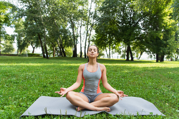młoda kobieta z zamkniętymi oczami siedząca w pozycji lotosu i medytująca na macie do jogi w parku  - Zdjęcie, obraz