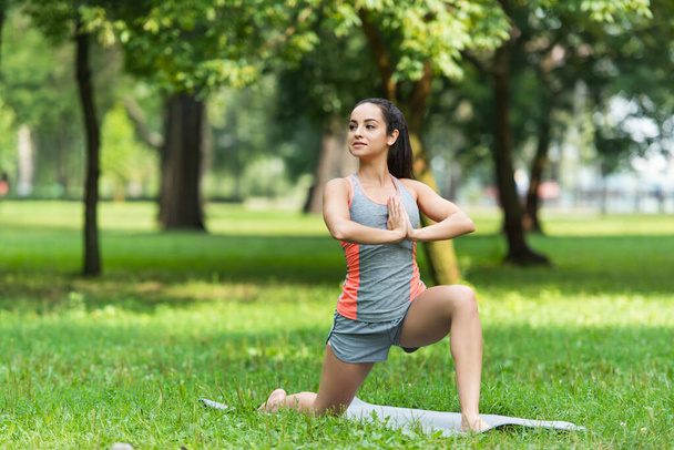 fit woman doing low lunge pose with praying hands on yoga mat in park - Foto, afbeelding