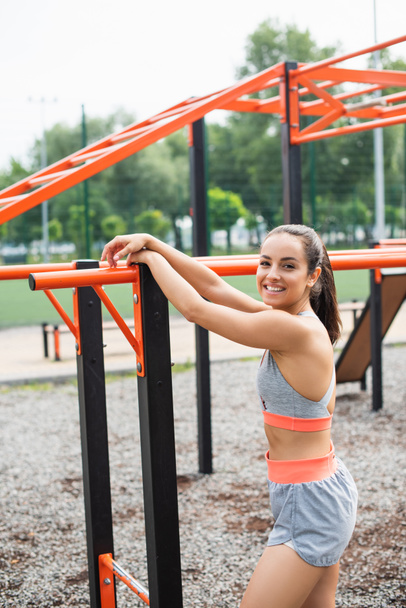 cheerful sportswoman smiling while leaning on parallel bars outside  - Fotoğraf, Görsel
