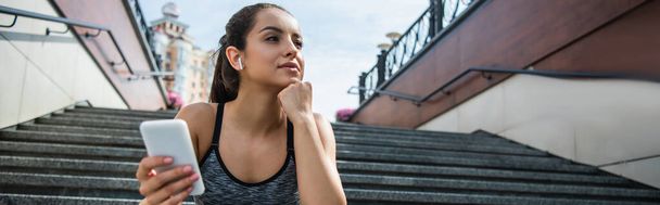 young sportswoman in wireless earphones using smartphone while listening music outside, banner - Foto, Imagen