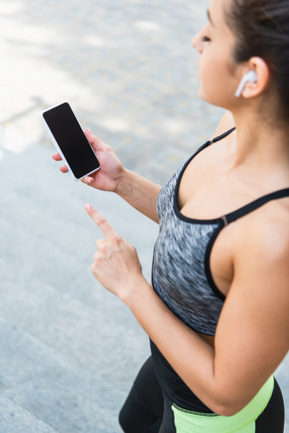 high angle view of sportswoman in wireless earphones using smartphone with blank screen outside  - Φωτογραφία, εικόνα