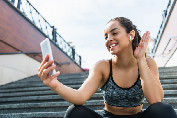 cheerful sportswoman in wireless earphones having video call on smartphone sitting on stairs outside  - 写真・画像