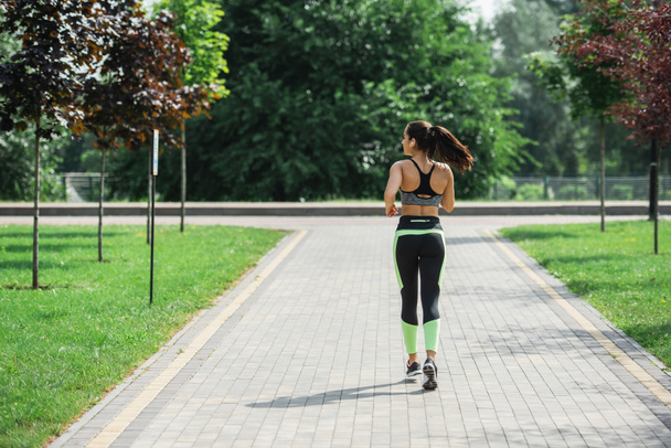 вид спортивной женщины в топе, леггинсы и беспроводные наушники, слушающей музыку во время бега в парке - Фото, изображение